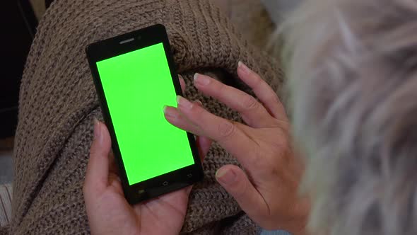 A Middle-aged Woman Sits and Works on a Smartphone with a Green Screen - She Scrolls Downward