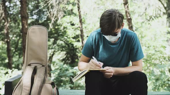 Young Man with a Medical Mask on Face Is Sitting on a Bench in Park.