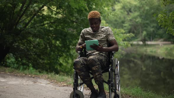 Smiling African American Man in Wheelchair Surfing Social Media on Tablet Sitting on Lake Shore in