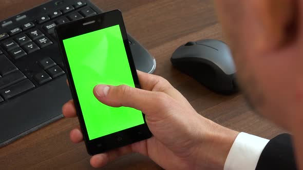 An office worker slides on a smartphone with a green screen at a desk with a computer - closeup