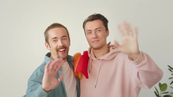Two Lgbt Men are Standing in the Living Room Looking at the Camera and Waving Hands