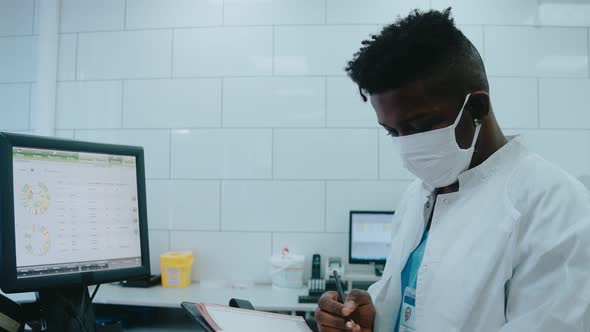 Black Doctor Writes Information From Computer in White Lab Wearing Mask and Gown