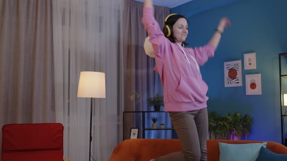 Happy Adult Woman in Headphones Dancing and Jumping While Listening Music at Home Alone on Sofa