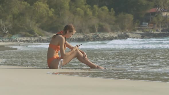 Girl in Orange Bikini with Phone Sits on Wet Sand