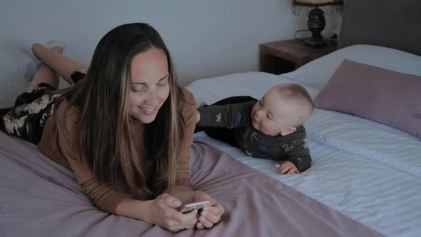 Mom and baby are lying on the bed. Young woman with her son using smartphone.