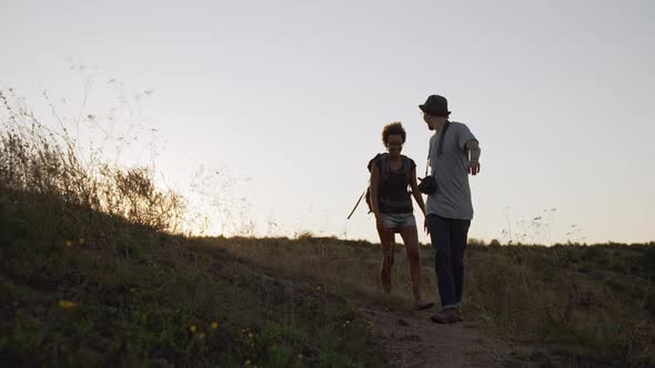 Caucasian Male with Camera Glasses and Hat Alking Outdoors with Darkskinned Female with Backpack in