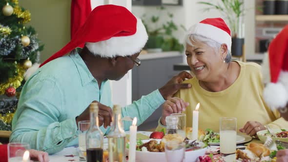 Happy diverse senior couple in santa hats talking at christmas dinner table at home