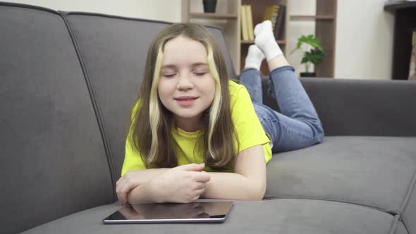 A Little Girl is Smiling While Lying on the Couch with a Tablet