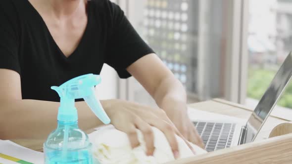 Young woman cleaning the surface of laptop with alcohol spray and fabric at home