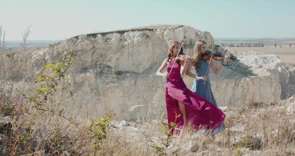 Two Women in Blowing Dresses Play Viola Among Cliffs in Sunny Summer