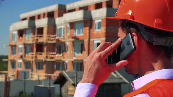 Construction Worker Phones with the Smartphone in Front of Building Site - Closeup From Back