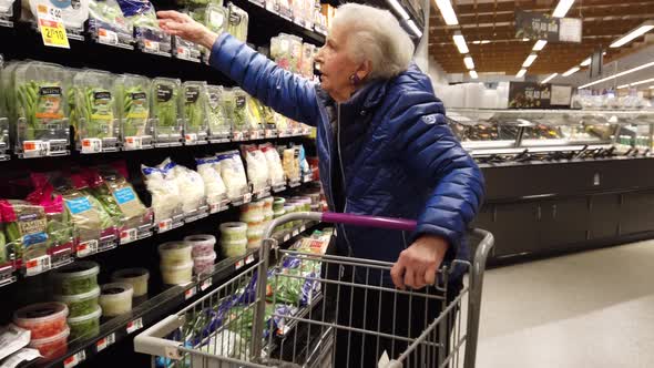 Elderly woman looking at bags of brussels sprouts in a grocery store.