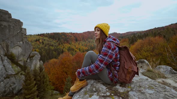 Woman Traveler in Casual Wear and Yellow Beanie with Backpack Sits on Edge of Cliff and Enjoing