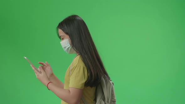 Side View Of Asian Girl Student Wearing A Mask And Use Tablet While Walking On Green Screen