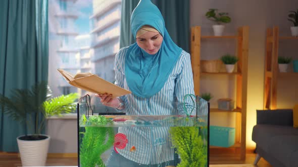 Muslim Woman in National Headscarf Studies a Book on Aquariums and Feeds Goldfish in an Aquarium