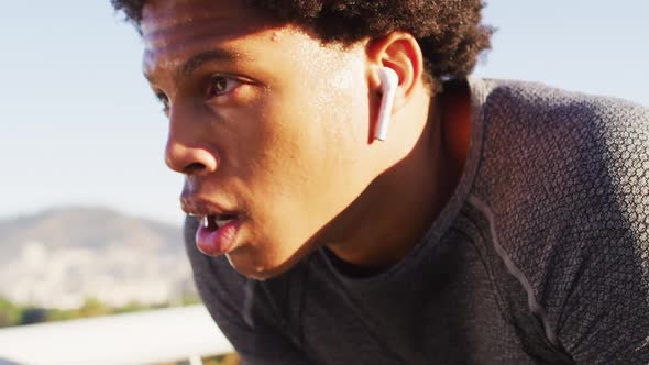 Fit african american man exercising outdoors in city, with wireless earphones, resting on footbridge