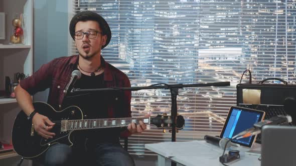 Guitarist in Hat and Trendy Casual Clothes Playing Guitar and Singing in Home Recording Studio