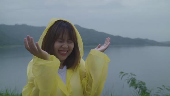 Young Asian woman feeling happy playing rain while wearing raincoat standing near lake.