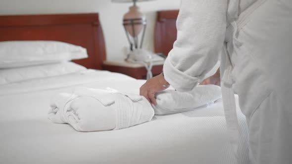 An Unrecognized Adult Senior Woman Puts a White Towel Next To a White Bathrobe on a Bed in a Hotel