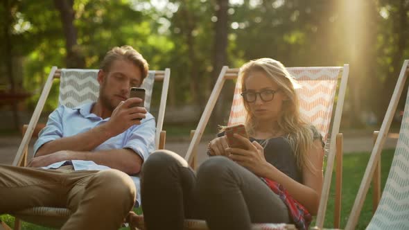 Caucasian Bearded Male and Blonde Female with Glasses Sitting on Lounges in Park with Mobile Phones