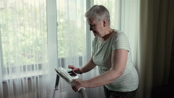 Old Woman Walks on a Treadmill in a Front of the Window