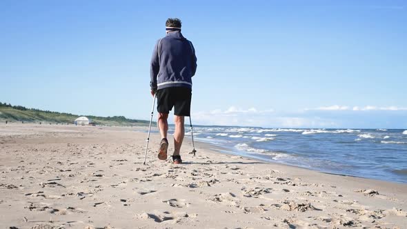 Old Man Uses Sticks for Scandinavian Walking on the Sea Beach, Sunny Day