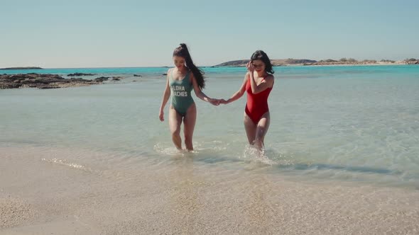 Pretty Female Friends Walking Holding Hands By Sea on Sandy Tropical Beach in Swimsuits