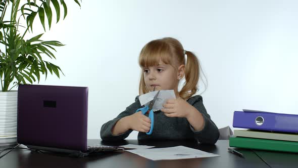 Child Schoolgirl Learns Lessons at Home Sitting at Table Cutting with Scissors Shapes Out of Paper