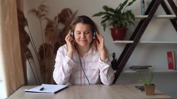 Young Beautiful Caucasian Woman Sits at Home at a Desk Listens to Music on Headphones and Dances