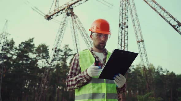 Electrical Equipment Worker Near High Voltage Tower Using Walkie Talkie and Clipboard