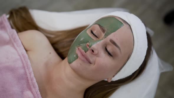 Girl Lying with Cosmetic Mask of Green Clay on Face Opens Eyes and Smiles
