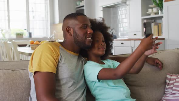 African american daughter and her father using smartphone together sitting on couch