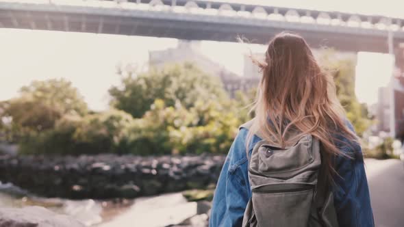 Camera Follows Young Female Tourist with Backpack Walking Along Sunny Brooklyn Park River Embankment