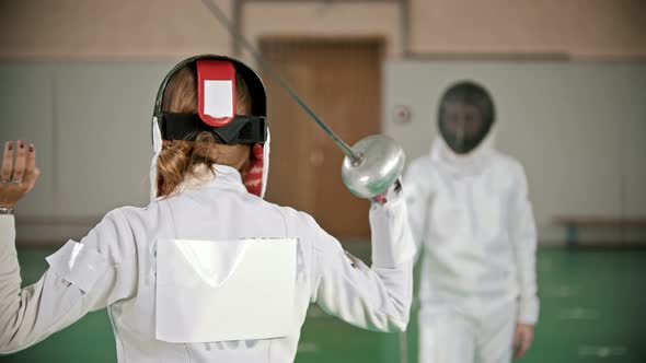 Two Young Women Fencers Having a Training in the Gym - a Woman Stretches with the Sword Behind Her