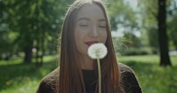 Young Spring Fashion Woman Blowing Dandelion in Spring Garden. Springtime. Trendy Girl at Sunset