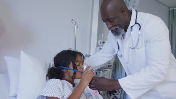 African american male doctor putting oxygen mask ventilator on mixed race girl lying in hospital bed