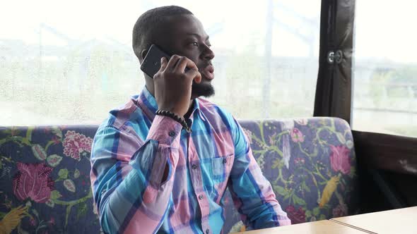 Afro American Man Calls and Talks Smartphone Sitting in Cafe Waiting His Order.