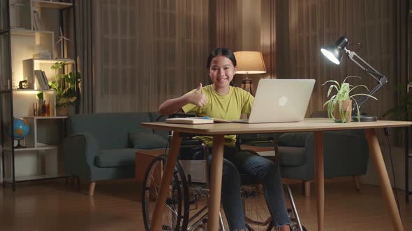 Asian Kid Girl Sitting In A Wheelchair Using Laptop Computer While Smiling And Thumb Up To Camera