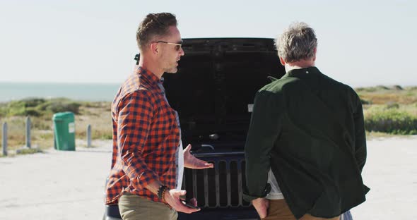 Stressed caucasian gay male couple standing by broken down car with open bonnet at the beach