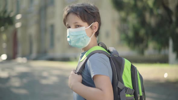 Sad Schoolboy with Backpack in Face Mask Turning To Camera. Portrait of Upset Caucasian Boy Standing