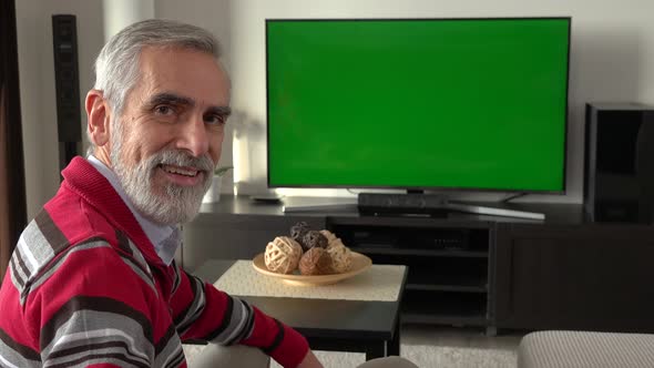 A Elderly Man Sits on a Sofa in Living Room and Watches TV with a Green Screen, Smiles at the Camera