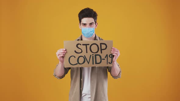 Young Man in Protective Mask is Raising Cardboard Poster with Inscription Stop Covid19 and Pointing