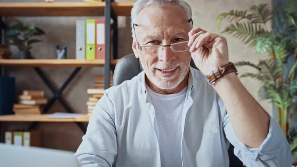 Senior Male is Sitting at Desk Next to His Notebook While Working at Home Office Putting on Glasses