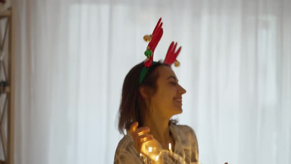 Happy Joyful Pretty Woman Wearing Reindeer Antlers Headband Dancing with Christmas Lights at Cozy