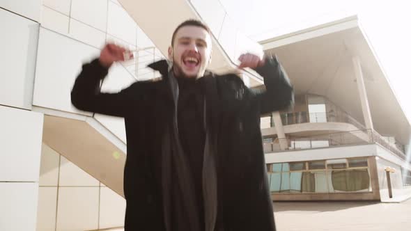 Young Handsome Caucasian Male in Black Coat Jumping for Joy and Smiling Outdoors