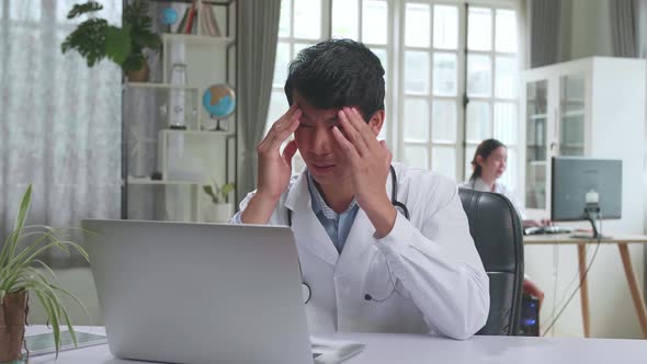 Young Asian Man Doctor Is Using Laptop Computer And Headache In Workplace. Medical Concept