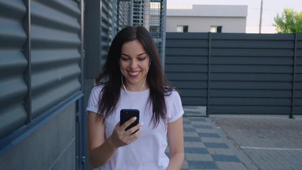 Portrait of Smiling Young Woman with Earphones Using Smartphone While Walking on the City Street