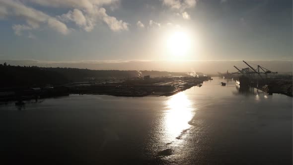 Bright golden sun reflecting off calm water, approaching an industrial port operation, aerial establ