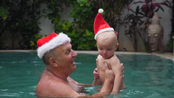 Happy Grandfather And Baby Wearing Santa Hats In Swimming Pool