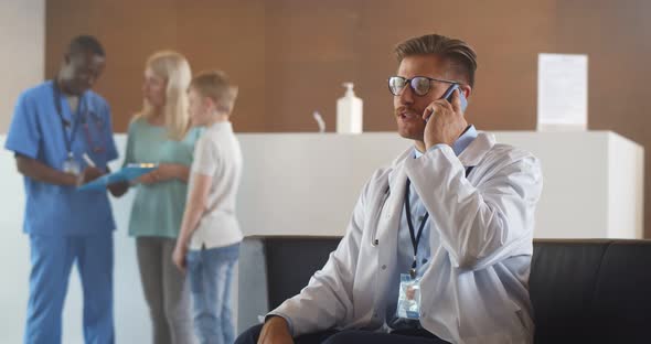 Young Male Doctor in White Coat Sitting on Sofa at Hospital Corridor Talking on Mobile Phone